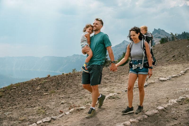 Parents hiking a mountain trail with their two young children, smiling and holding hands with mountains in the background