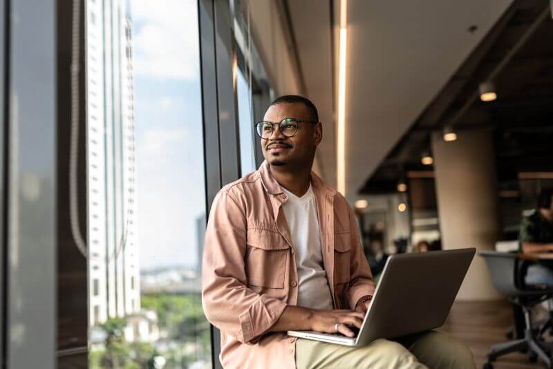 Man with a laptop looking out a city office window with a confident smile, weighing his home equity and personal loan options before making the right borrowing decision