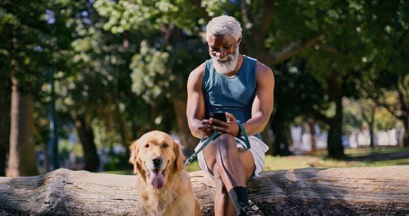 Man sitting on a log in the park with his dog, checking personal loan rates on his phone