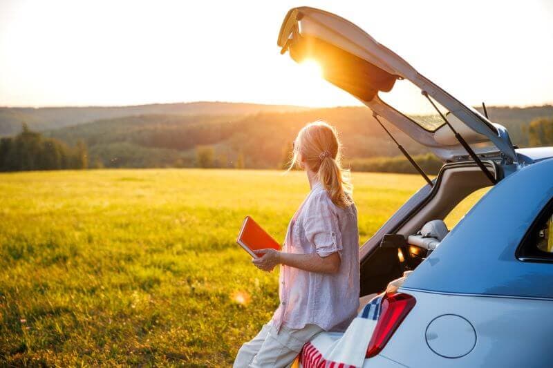 Woman sitting on car trunk in a sunny field, holding a notebook while considering her debt payoff plan